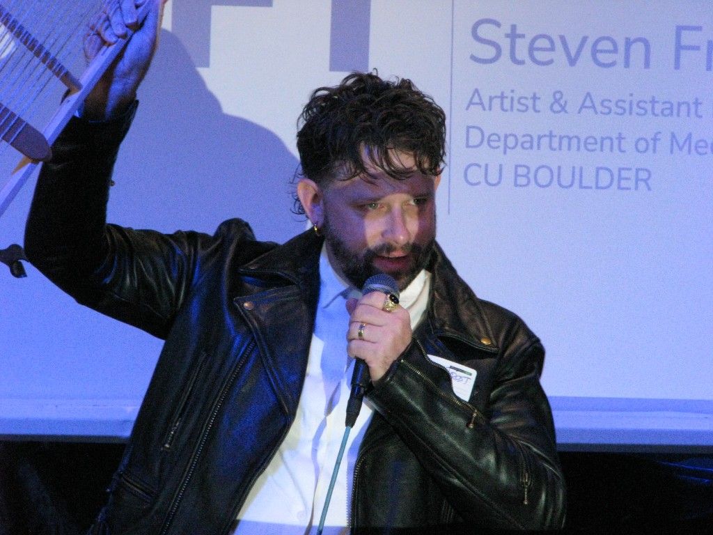 Speaker in a leather jacket holding up a string instrument while talking into a microphone