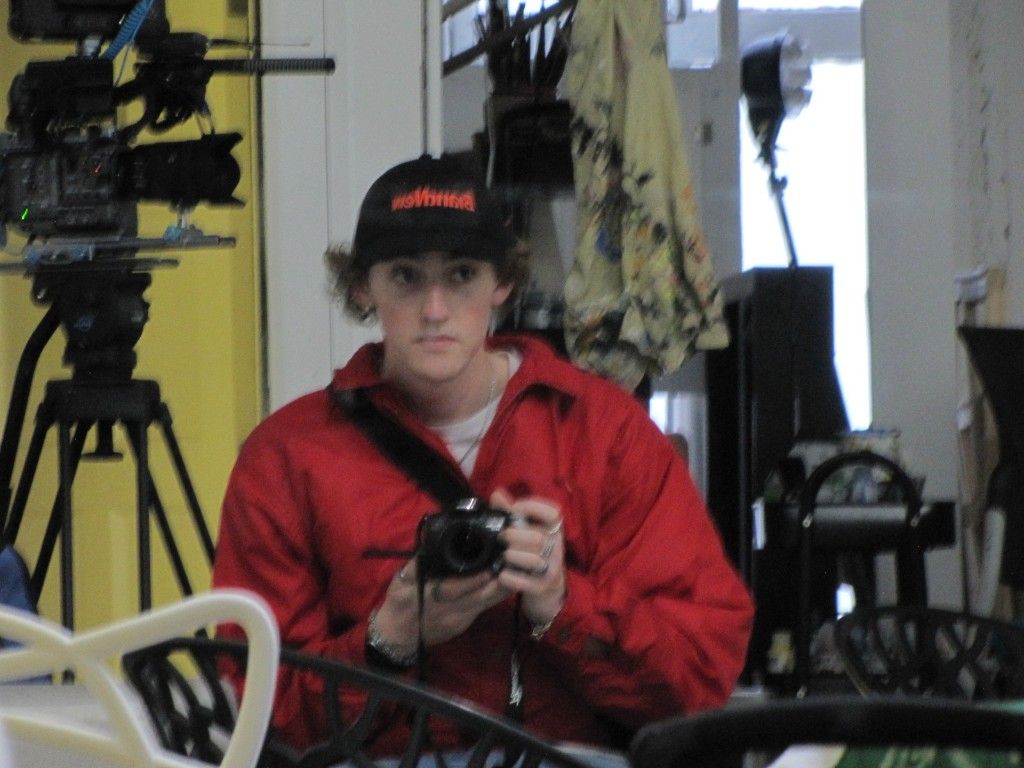 Kai in a red jacket holding a camera, reflected in a mirror beside a film camera rig