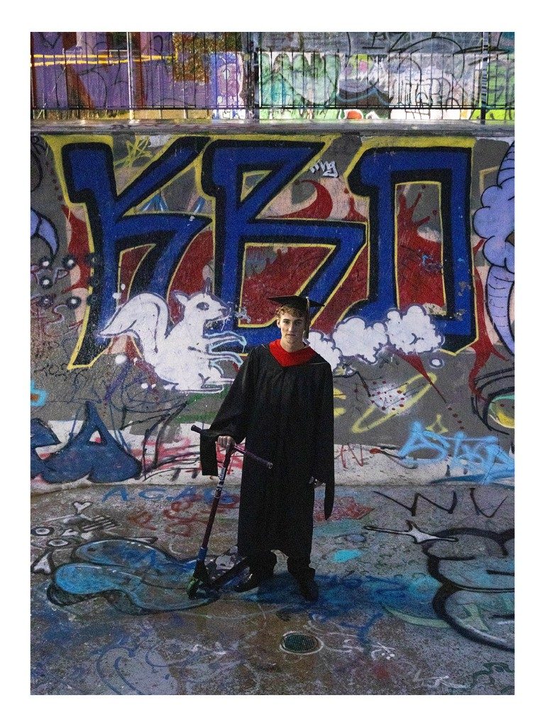 Graduate standing with a scooter in front of a graffiti wall