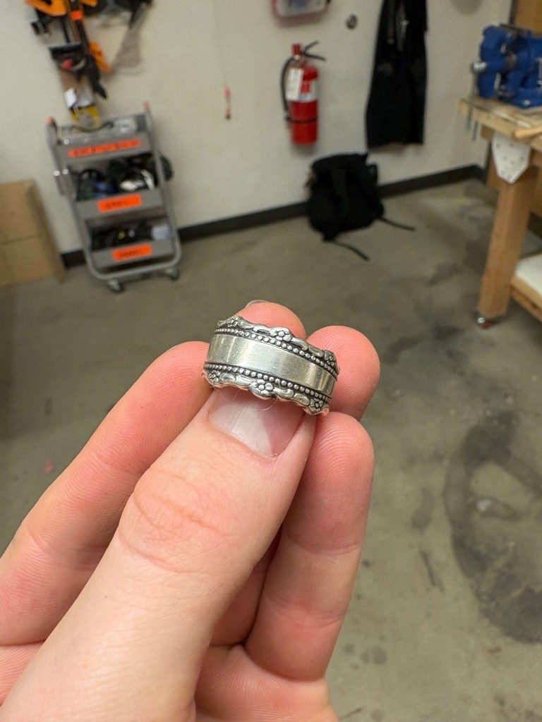 Beaded-edge silver spoon ring held between fingers in workshop